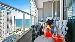 Un balcón con vista a una playa y edificios altos, una mesa con bebidas y fruta, y una silla.