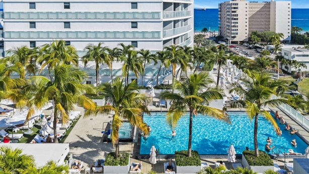 Un resort con piscina central rodeada de palmeras, sillas de descanso y vista al océano.