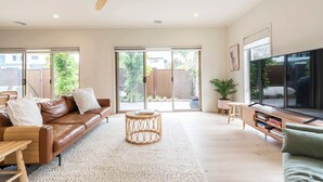 Een moderne woonkamer met een bruine leren bank, een ronde rieten salontafel, een houten tv-meubel en een groot raam met uitzicht op groen.