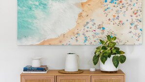 Een kunstwerk met een strandthema, een bloempot, boeken en een rieten mandje op een houten kast.