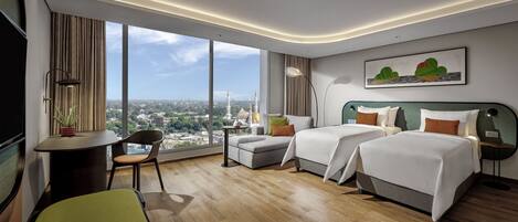 A hotel room with two beds, a desk, a chair, and a large window offering a city view.