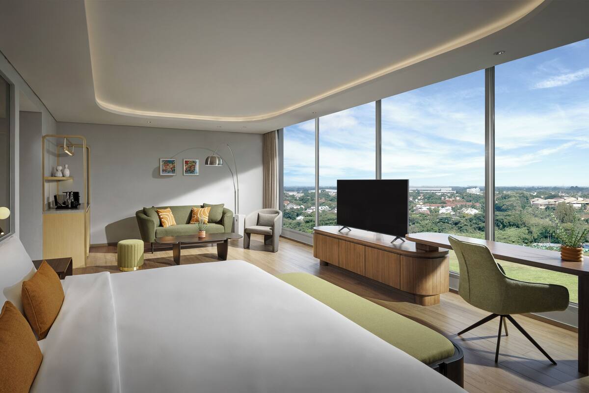 Une chambre d’hôtel moderne dotée d’un grand lit, d’un canapé, d’un bureau avec une télévision et offrant une vue sur la ville grâce à de grandes fenêtres.