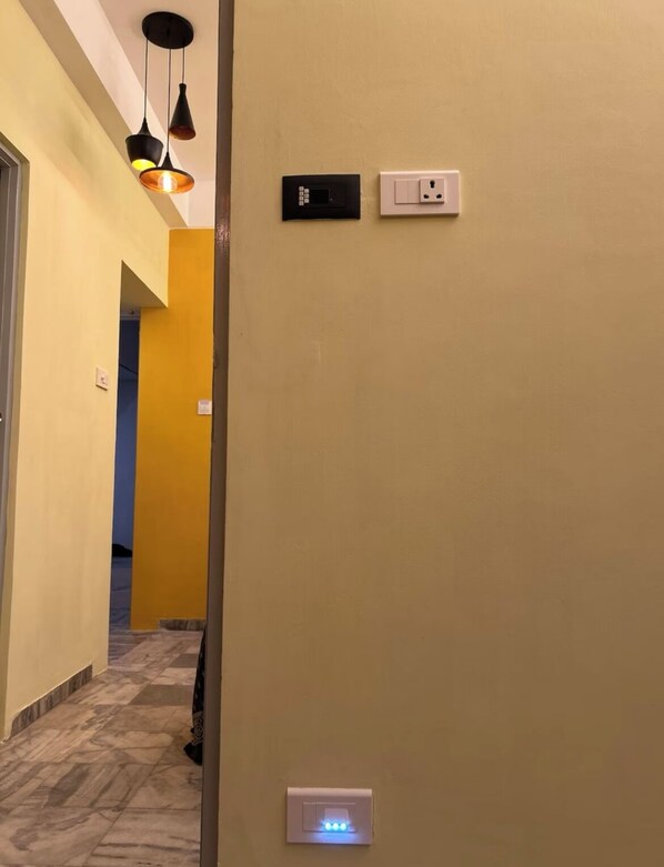 Un pasillo con pared amarilla, un portero automático negro, un interruptor de luz y un botón azul iluminado.