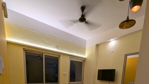 A room with a yellow sofa set, a small table, a ceiling fan, and a TV mounted on the wall.