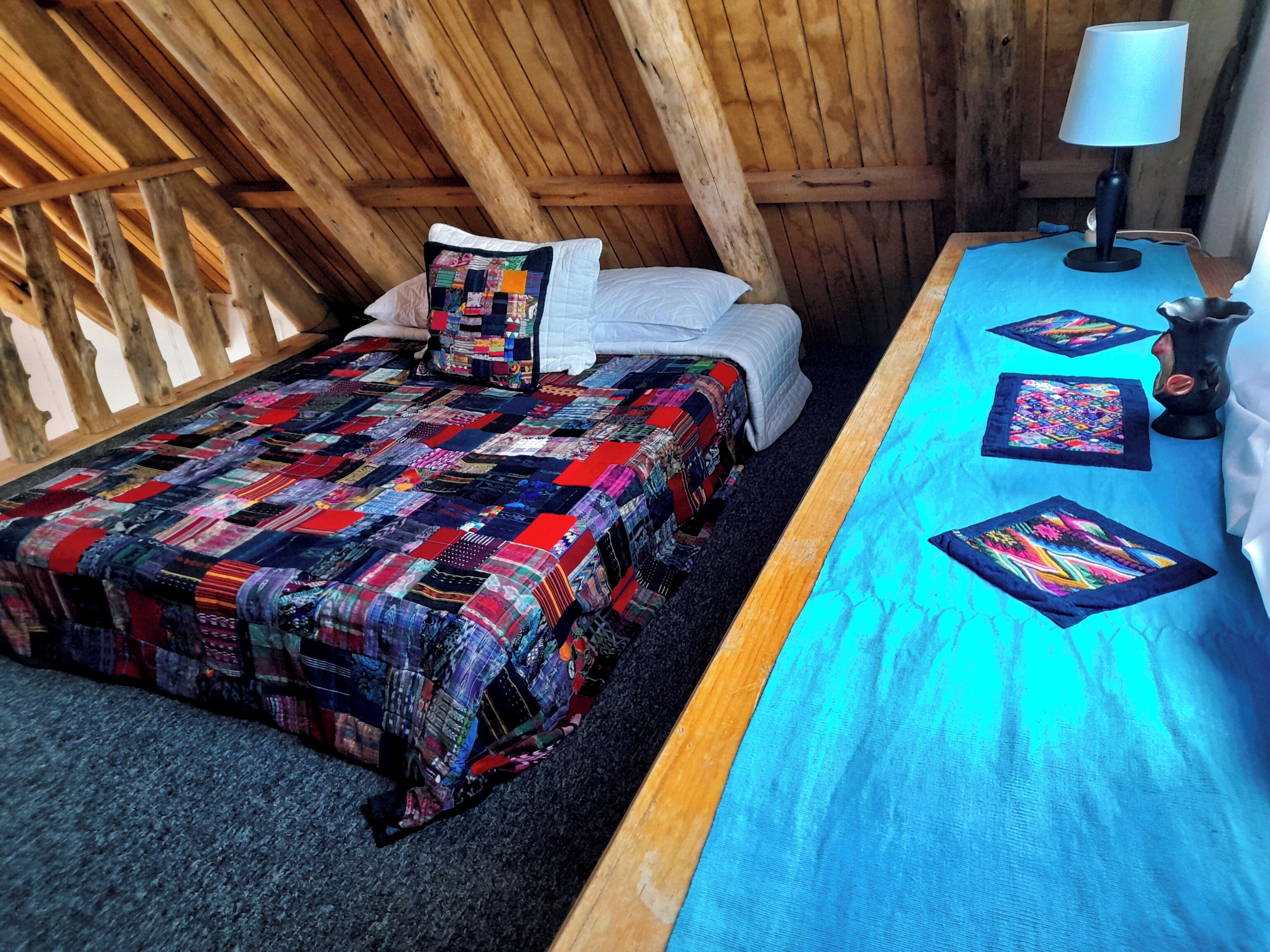 A cozy room with a wooden ceiling, a bed with a colorful quilt, a blue table with a lamp, and a small decorative item.