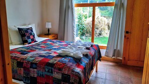 A bedroom with a large bed, a nightstand with a lamp, and a view of a garden through a window. - El Colibri Cabin (San Juan La Laguna)