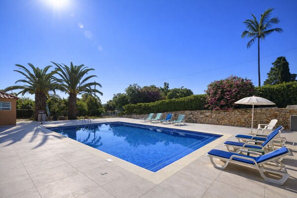 Une piscine rectangulaire à l’eau d’un bleu limpide, entourée d’une terrasse carrelée. On y trouve des chaises longues avec des coussins bleus, ainsi qu’un mur de pierre agrémenté d’un petit jardin et d’un palmier.
