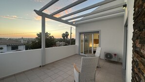 Een balkon met uitzicht op de ondergaande zon, een witte stoel en een schuifdeur naar een slaapkamer.