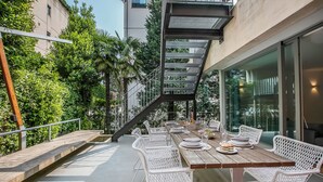 Un espace de restauration en plein air avec une longue table en bois, des chaises en osier blanches et un escalier menant à un niveau supérieur.