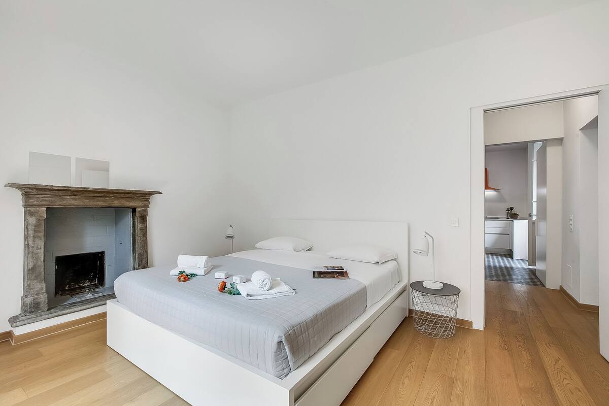 A modern bedroom with a bed, a fireplace, a bedside table, and a hallway leading to a kitchen.