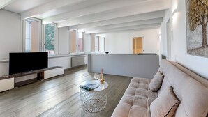 Un salon moderne avec un téléviseur à écran plat, une table basse ronde sur laquelle se trouvent un livre et des lunettes, un canapé beige et une grande fenêtre donnant sur des arbres.