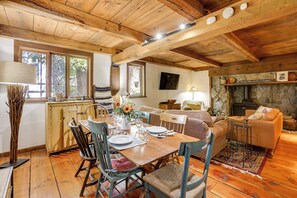 Un'accogliente stanza in legno dallo stile rustico, con un tavolo da pranzo apparecchiato per quattro persone, circondato da sedie. È presente un camino in pietra con un divano e poltrone nelle vicinanze.
