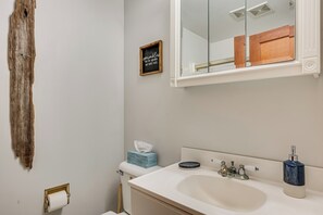 Ein Badezimmer mit Waschbecken, Spiegel und Toilette. An der Wand hängt ein Holzschild und dort ist eine Toilettenpapierrolle angebracht.