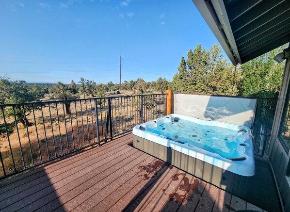 A hot tub on a wooden deck with a metal railing, overlooking a dry landscape with trees and power lines.