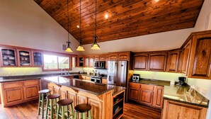 Una cocina con armarios de madera, una isla grande con taburetes, electrodomésticos de acero inoxidable y techo de madera.