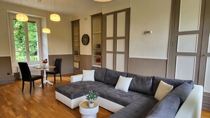 A modern living room with a sectional sofa, a dining area with a table and chairs, and a view of greenery through large windows. - Gîte du Château - 18 people - Centre de la France - au coeur du Berry (Cluis)