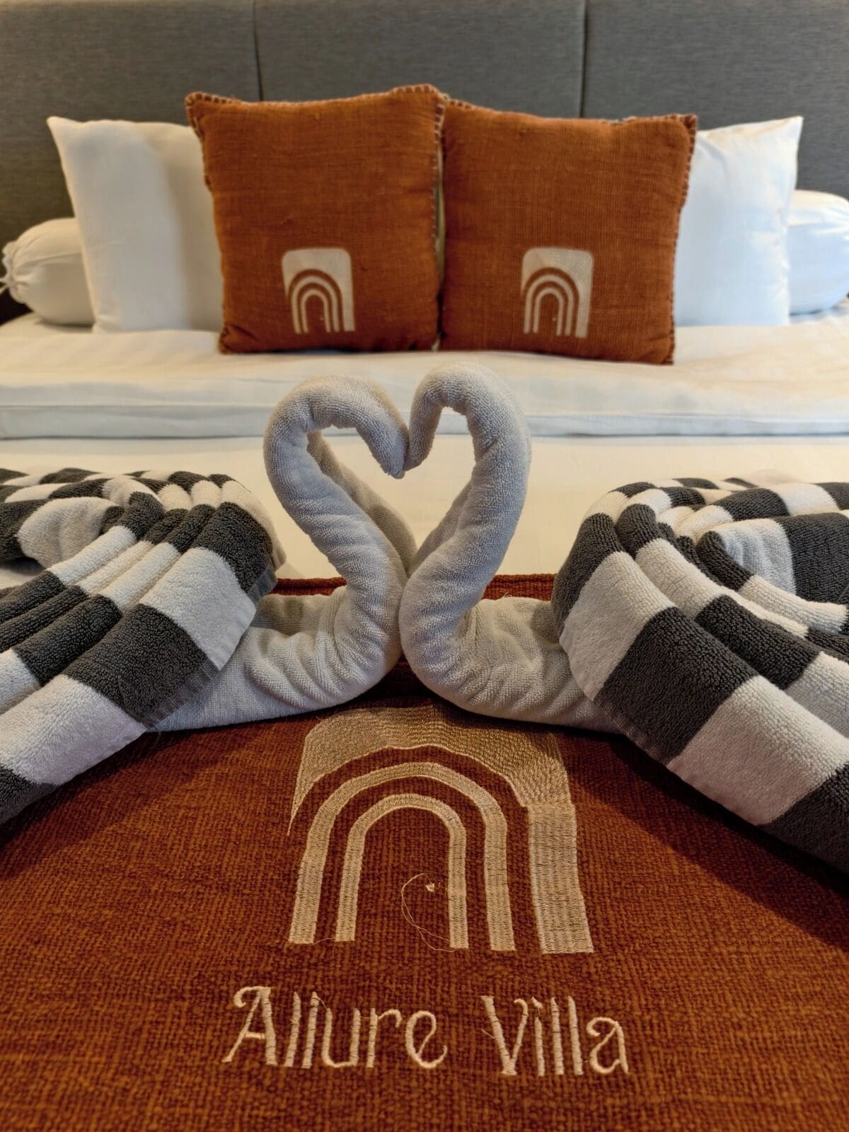 A bed with two pillows, a heart-shaped towel arrangement, and a towel with "Allure Villa" embroidered on it.