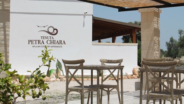 An outdoor seating area with wicker chairs and a white wall displaying the logo and name of Tenuta Petra Chiara. - Tenuta Petra Chiara -Apulian Country B&B (Nardo)
