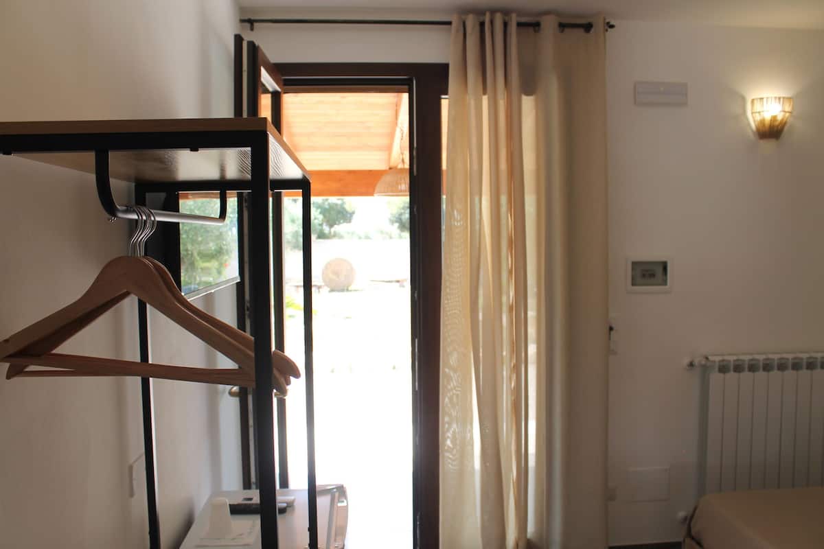 A hotel room with a bed, a radiator, a wall-mounted light, a door with curtains, and a wooden hanger on a wall.