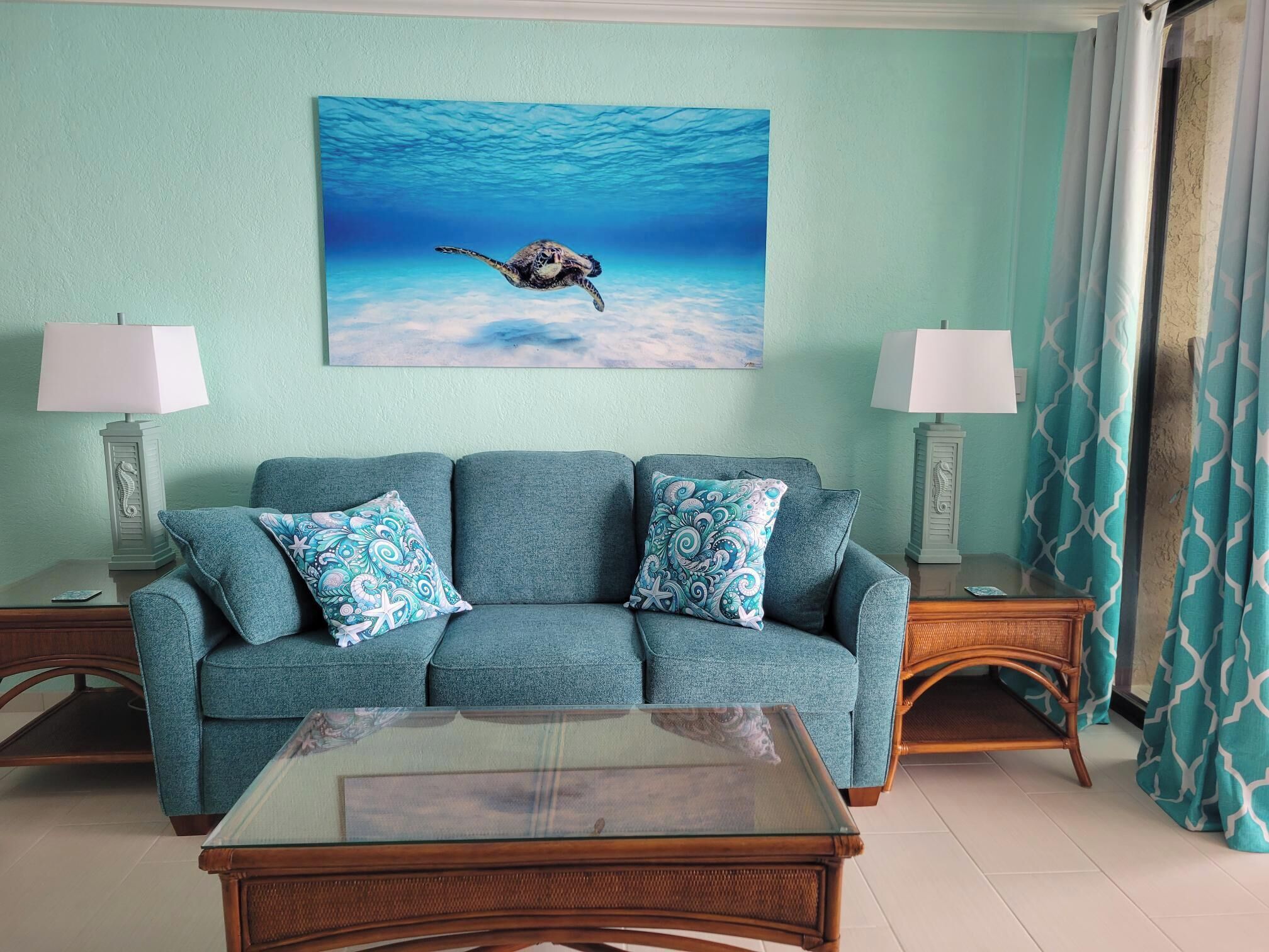 Ein Wohnzimmer mit einem türkisfarbenen Sofa, gemusterten Kissen, einem Glastisch und einem Gemälde einer Meeresschildkröte.