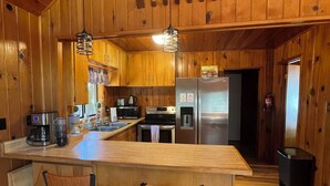 A wooden cabin interior with a kitchen, two bar stools, a refrigerator, and a hanging light fixture.