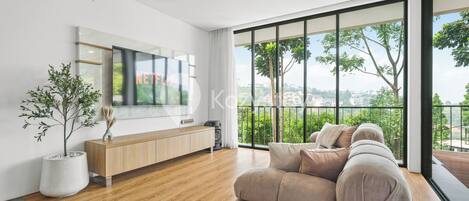 Una sala de estar moderna con un televisor grande, piso de madera, un sofá color beige, una consola de madera y un ventanal con vistas a zonas verdes y edificios.