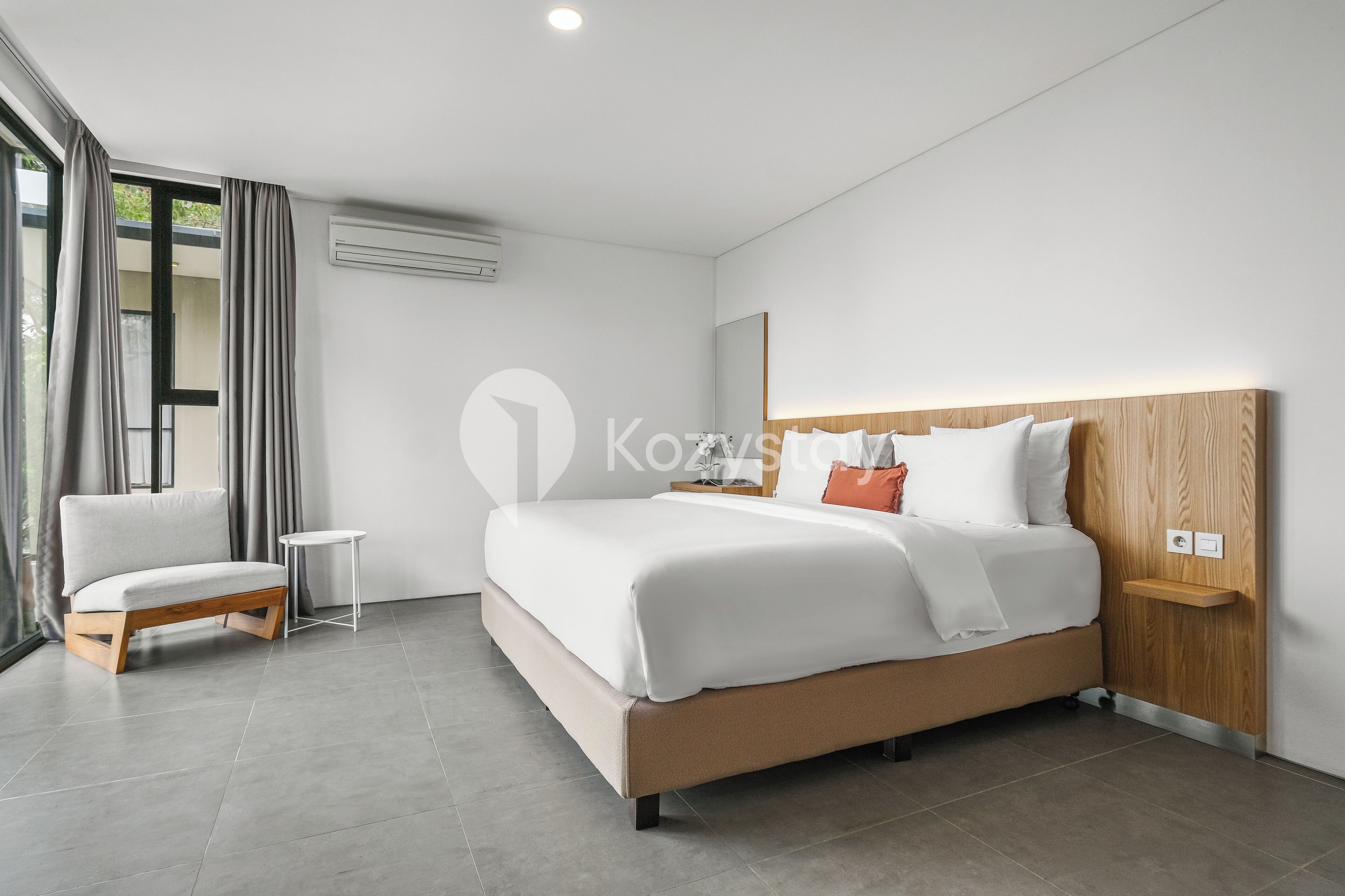 A modern hotel room with a large bed, a small bedside table, a chair, and a large window with curtains.