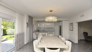 En moderne spisestue med et stort bord og stole, en lysekrone og udsigt til en have gennem en skydedør.