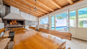 A wooden dining table with benches, a fireplace, and a view of a swimming pool through large windows.