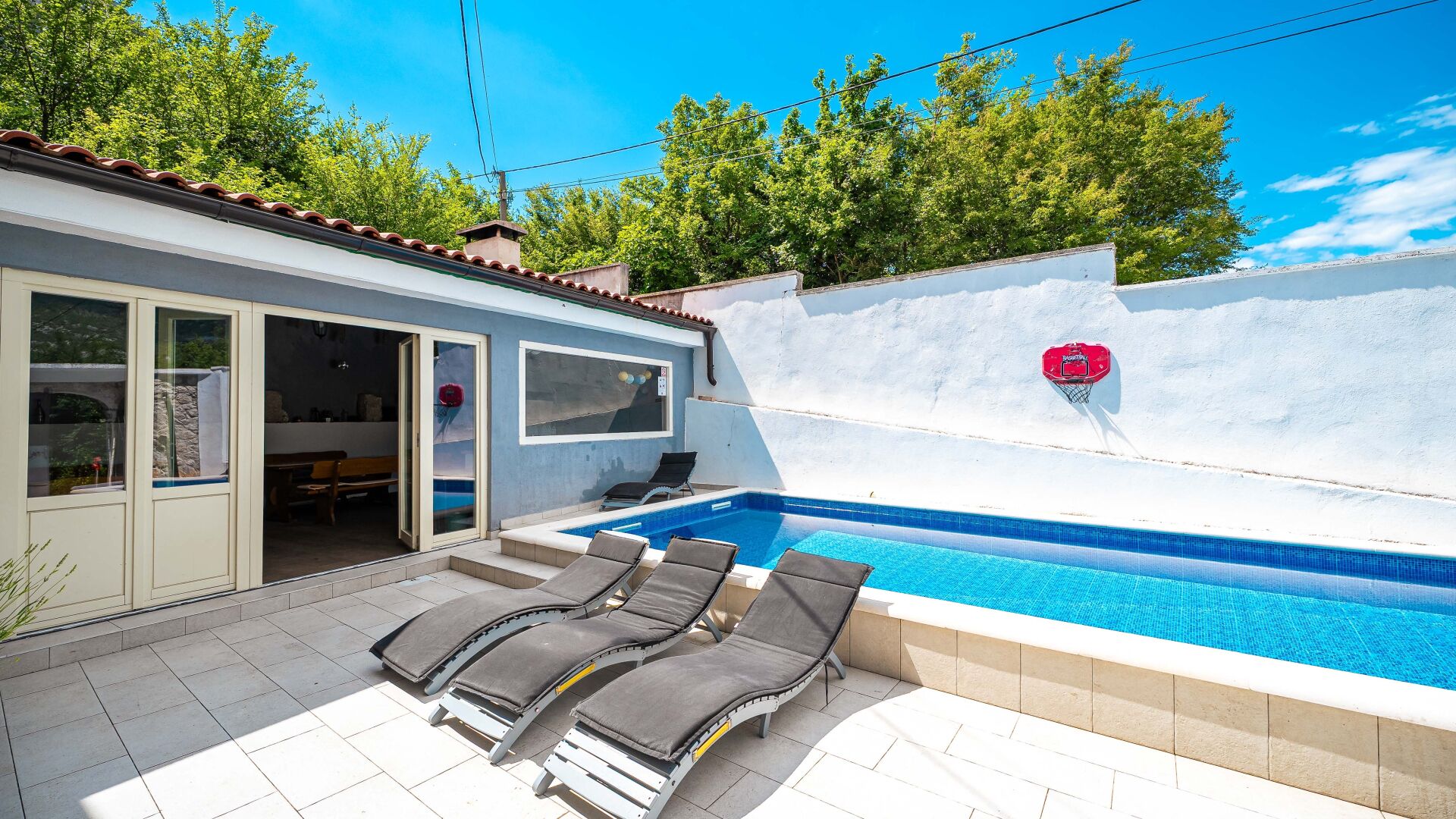 A swimming pool with a basketball hoop, lounge chairs, and a covered patio area.