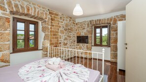 A bedroom with stone walls, a bed with floral bedding, a TV, and two windows with views.