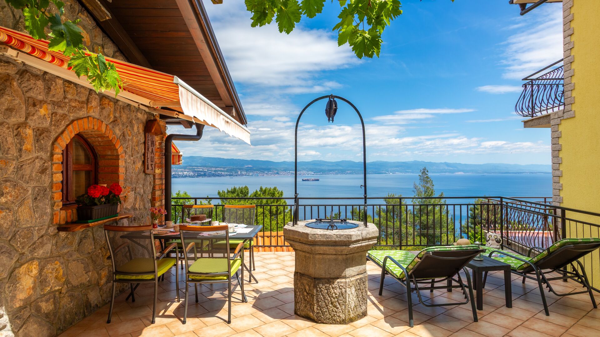 A terrace with stone walls, a table with chairs, a stone fountain, and a view of the sea.