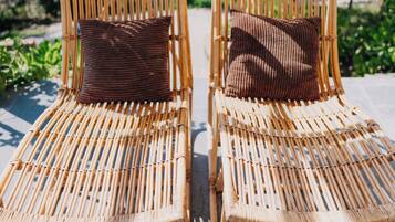 Twee rieten loungestoelen met kussens, geplaatst op een betegeld terras met uitzicht op groen en een gebouw op de achtergrond.