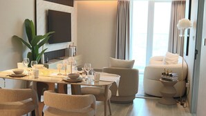 A modern living room with a dining area, a flat-screen TV mounted on the wall, a large plant, and a sofa with decorative items. - Condor Concept 7 - JVC Holiday Homes (Dubai)
