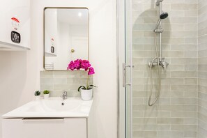 Un baño con un lavamanos blanco, un espejo, una orquídea en maceta y una ducha con mampara de vidrio y cabezal de ducha manual.