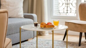 A cozy living room with a round table, a grey sofa, and a window with a balcony view. - Family Suites - Martin III-IV (Paris)