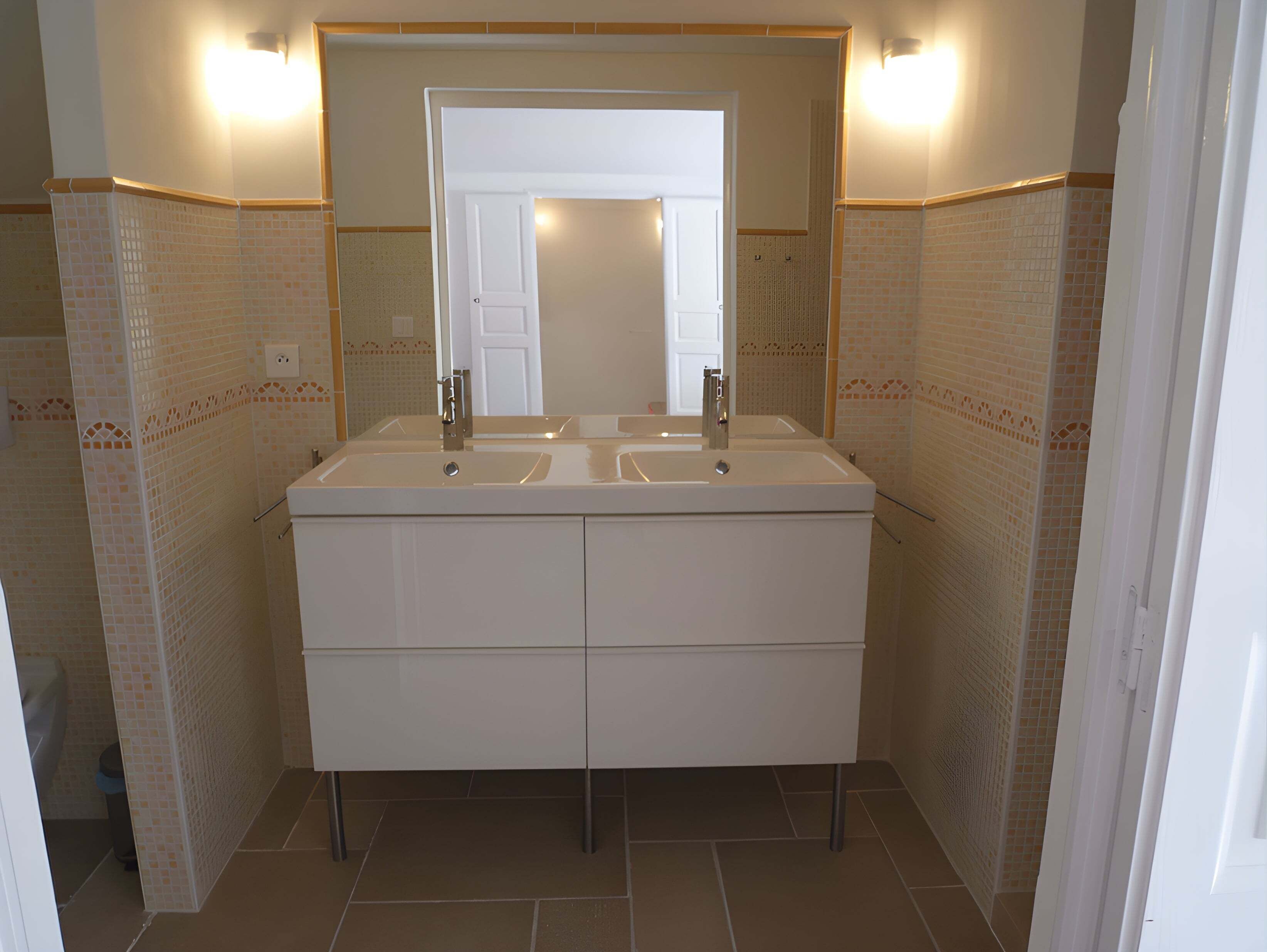 Een badkamer met een wastafelmeubel met dubbele wastafel, een grote spiegel en betegelde wanden.