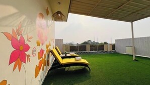 Eine Dachterrasse mit Kunstgrastrittichkeit, einem gelben Liegestuhl und einer Wand mit floralen Kunstwerken.