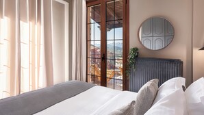 A bedroom with a bed, curtains, a window with a view, a radiator, and a round mirror on the wall. - Tilemachos by GM Luxury Suites Kalavryta (Καλαβρύτa)