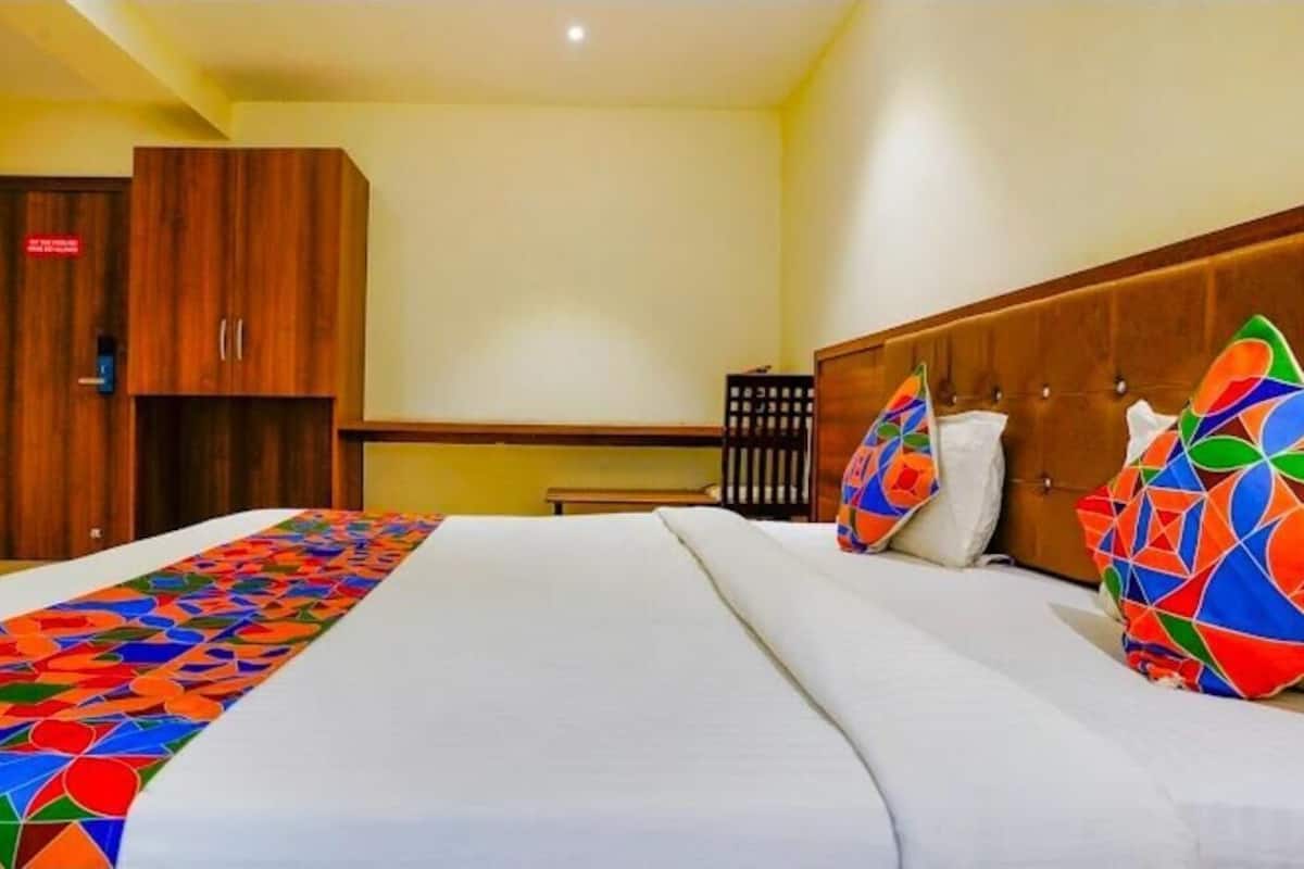 A hotel room with a bed, a wooden wardrobe, a bench, and a colorful decorative pillow.
