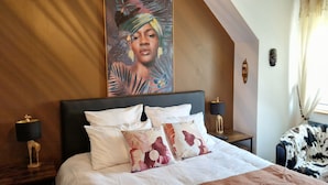 Een stijlvol ingerichte hotelkamer met een groot bed, meerdere kussens, een nachtkastje met lamp en een decoratieve wandhanger, plus uitzicht op het buiten.