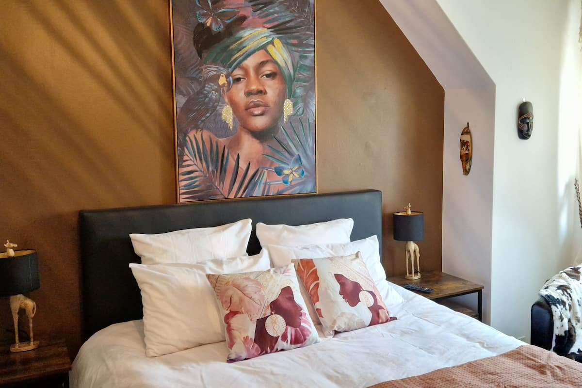 Una habitación de hotel elegante con una cama grande, varias almohadas, una mesita de noche con lámpara, un adorno de pared y vistas al exterior.