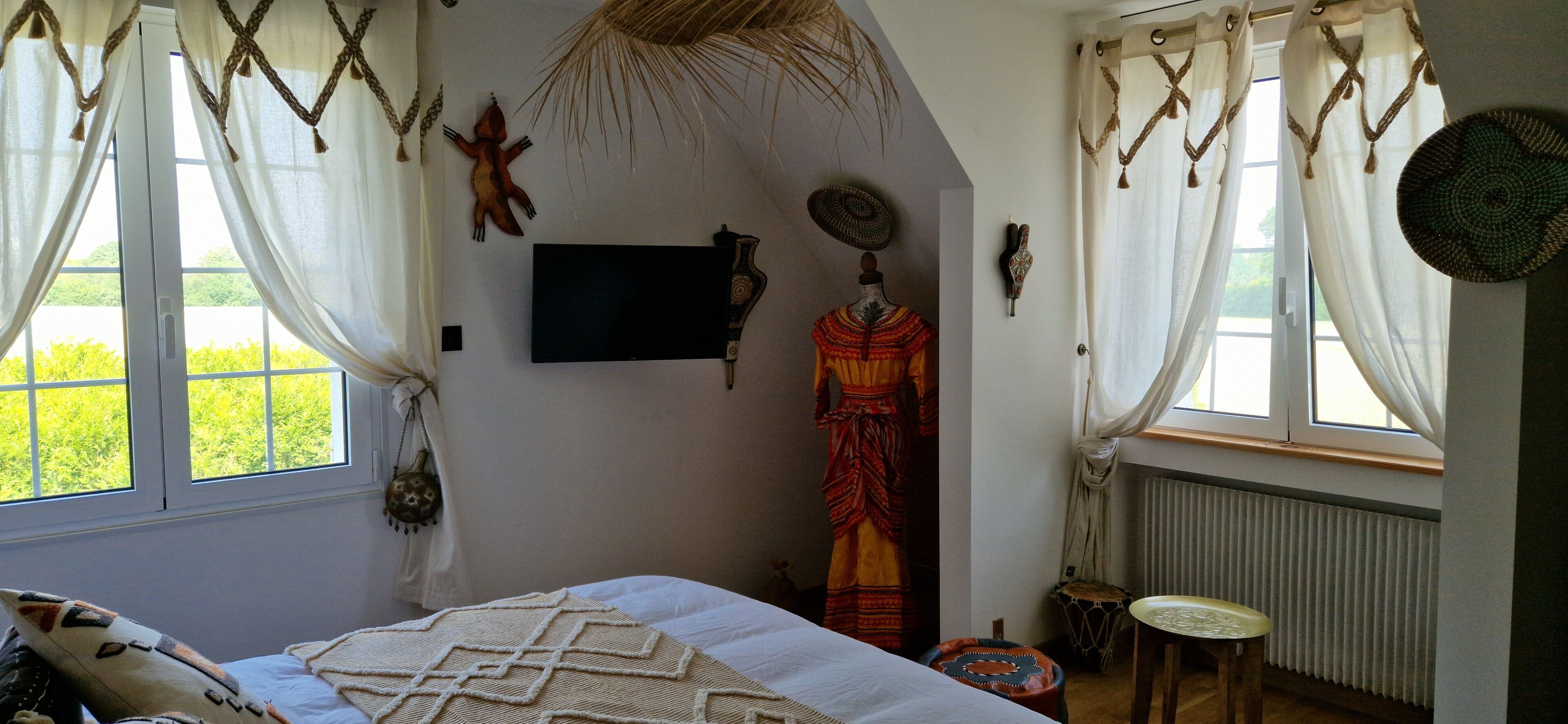 Une chambre avec un lit, une fenêtre avec des rideaux, un téléviseur fixé au mur et un mannequin vêtu d’un costume traditionnel.