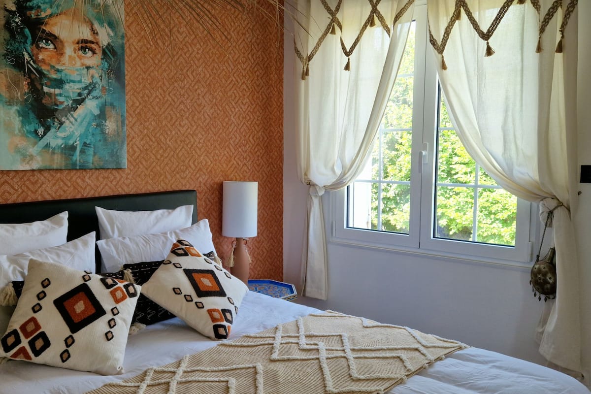 Un dormitorio con cama, almohadas estampadas, un cuadro, una lámpara y una ventana con cortinas.