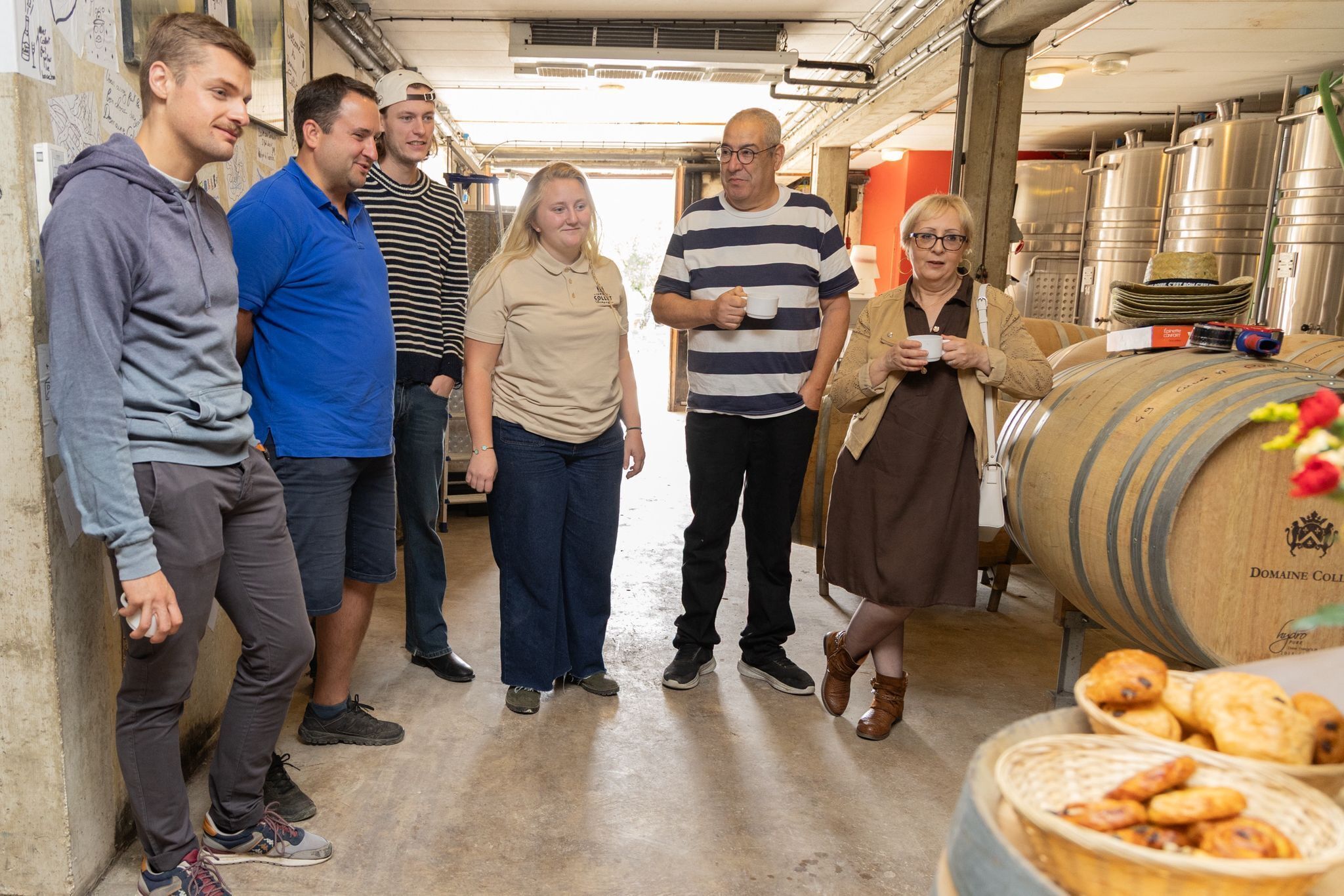 Eine Gruppe von Menschen in einem Raum mit Edelstahlbehältern und einem Holzbottich, vermutlich eine Weinkellerei oder Brauerei.