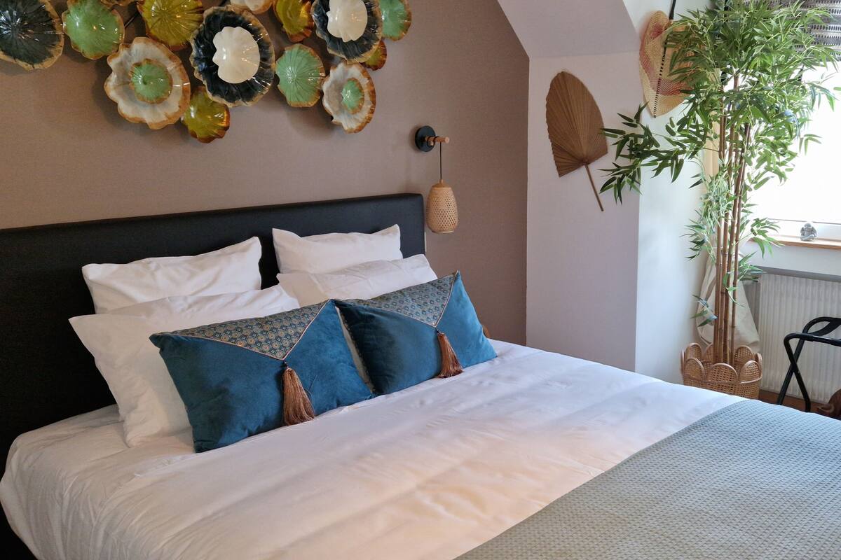 Una cama bien hecha con ropa de cama blanca y azul, una pared decorativa con arte de vidrio, una planta de bambú y una ventana con cortinas.