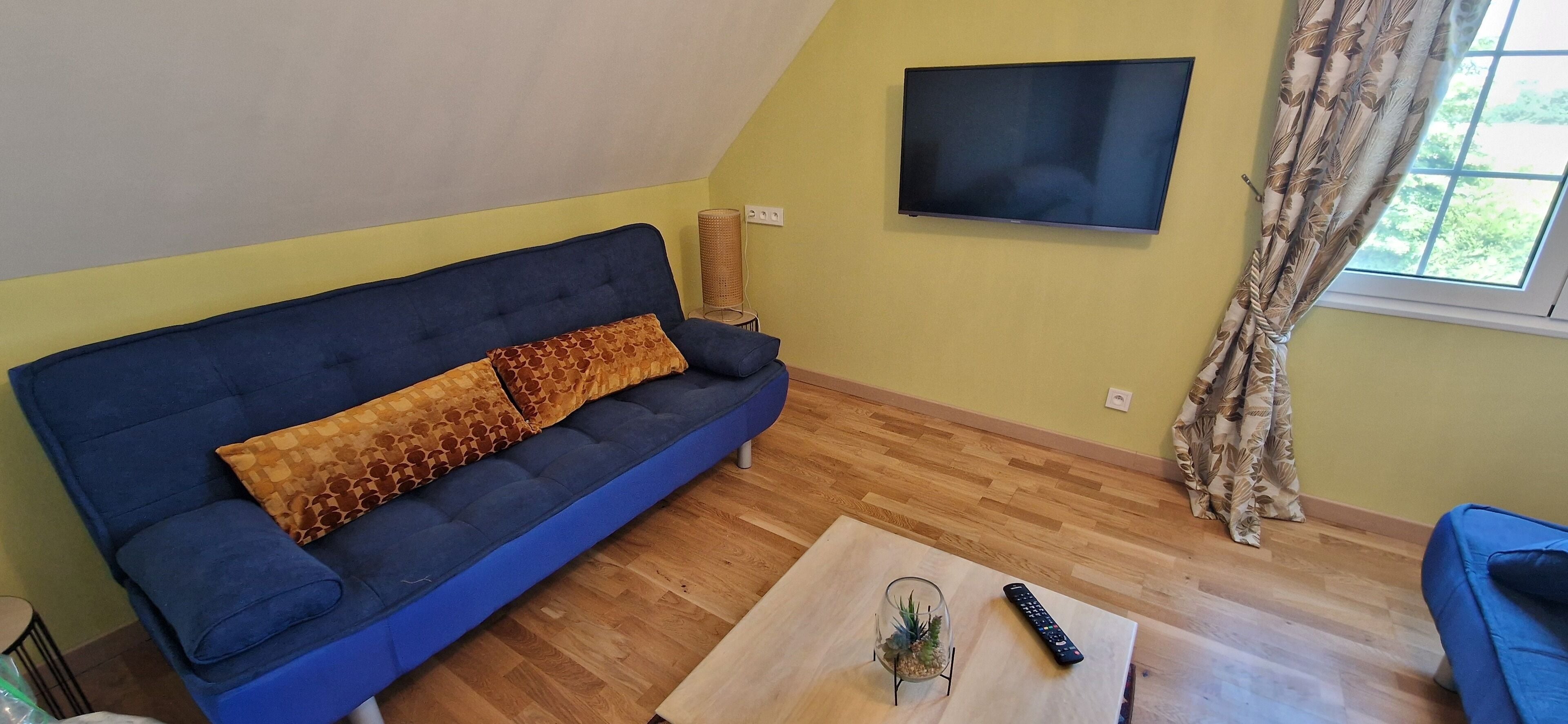 A living room with a blue sofa, a glass coffee table, a remote control, and a TV on the wall.