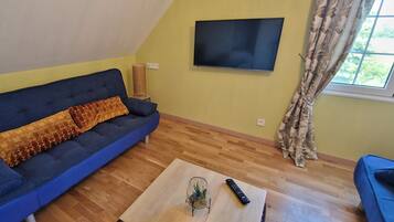 A living room with a blue sofa, a glass coffee table, a remote control, and a TV on the wall.