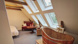 A room with a sloped ceiling, wooden beams, a large bathtub, a sofa, a small table with candles, and a view through skylights. - Aamølle Gæstgiveri (Hadsund)