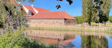 En byggnad av sten med ett rött tak, en damm och omgivande grönska.